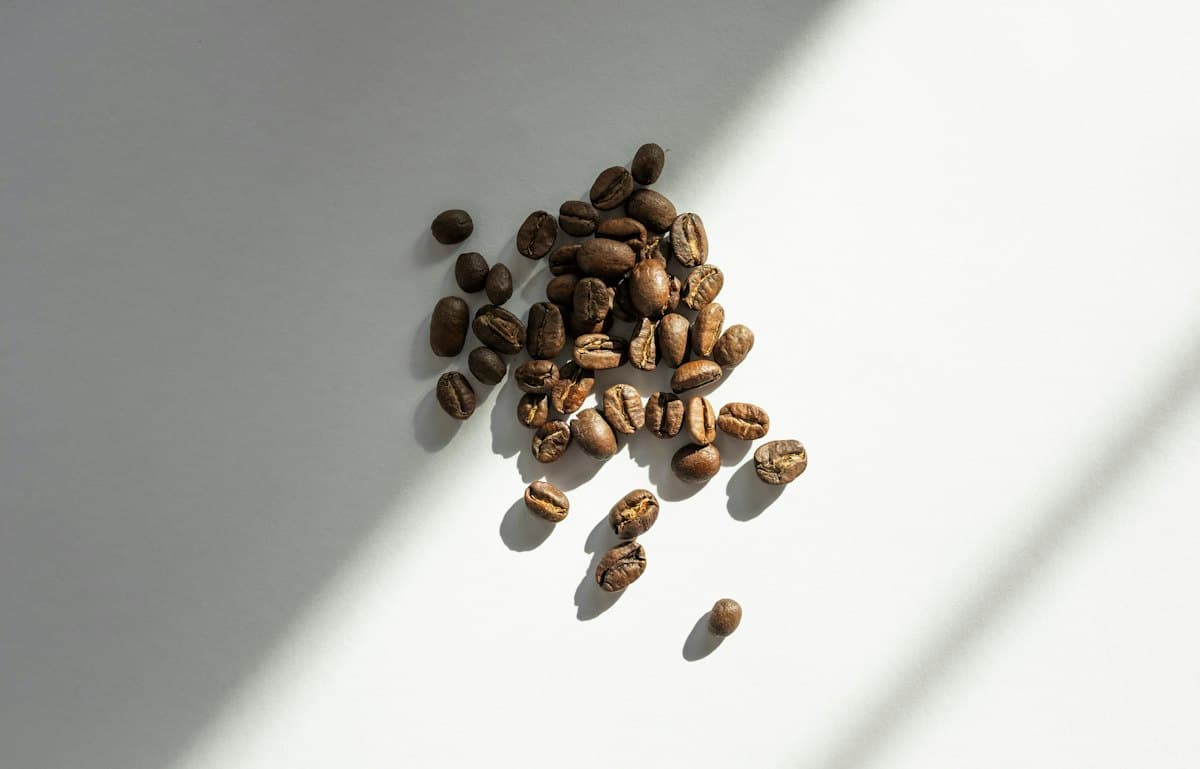 Boabe de cafea prăjite, risipite pe o suprafață albă, în lumină naturală