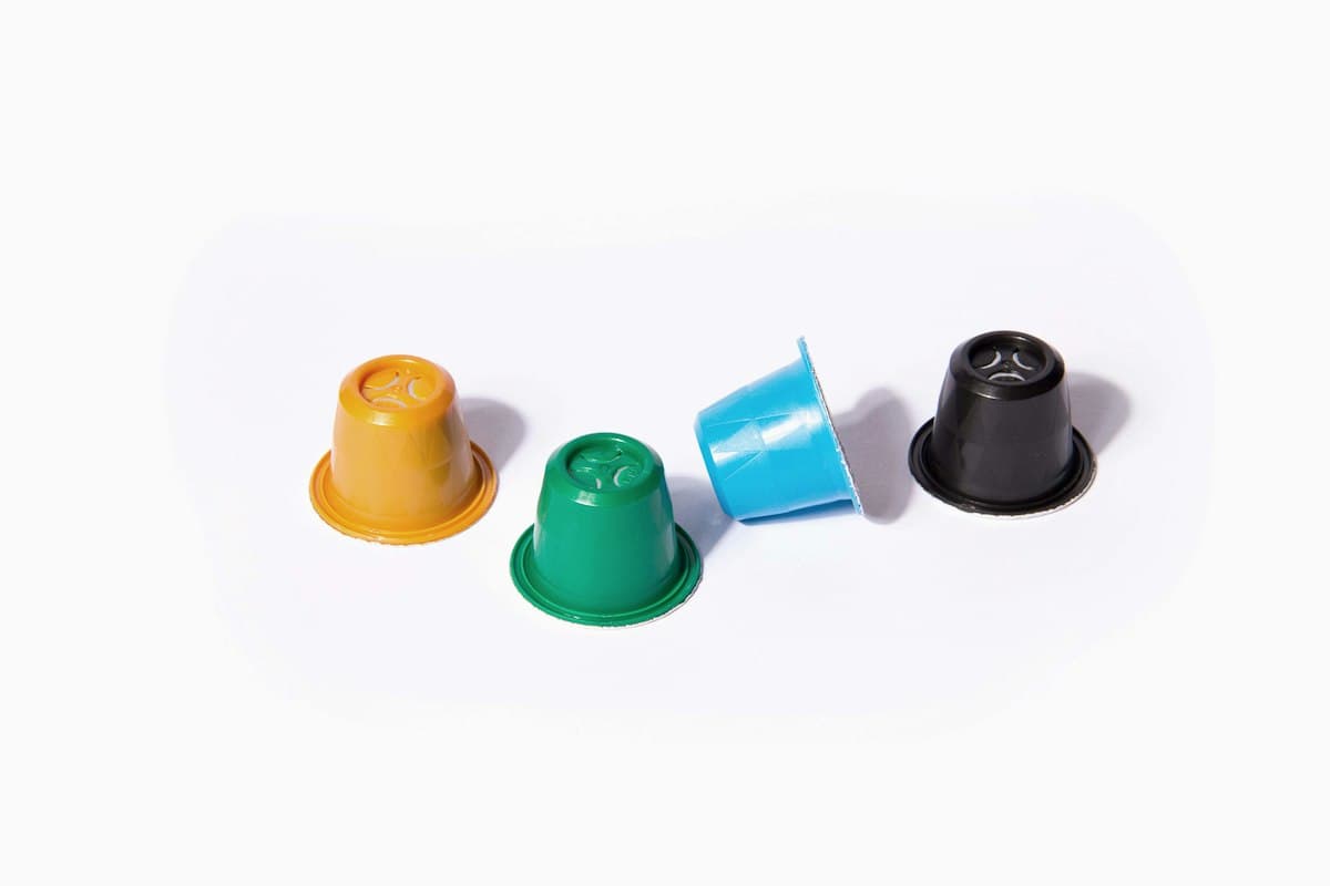 Patru capsule de cafea colorate, galbenă, verde, albastră și neagră, pe fundal alb